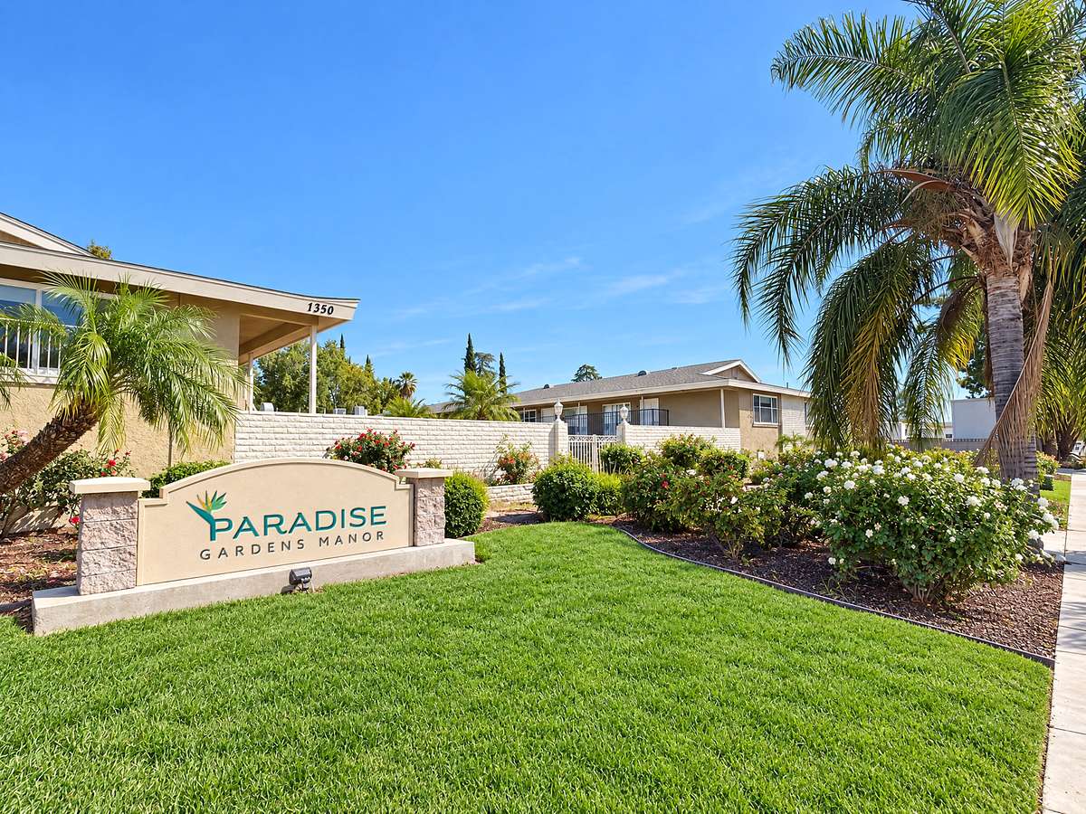 The front of Paradise Gardens Manor, with the Paradise Gardens Manor sign on its stone base, flowers and palm trees
