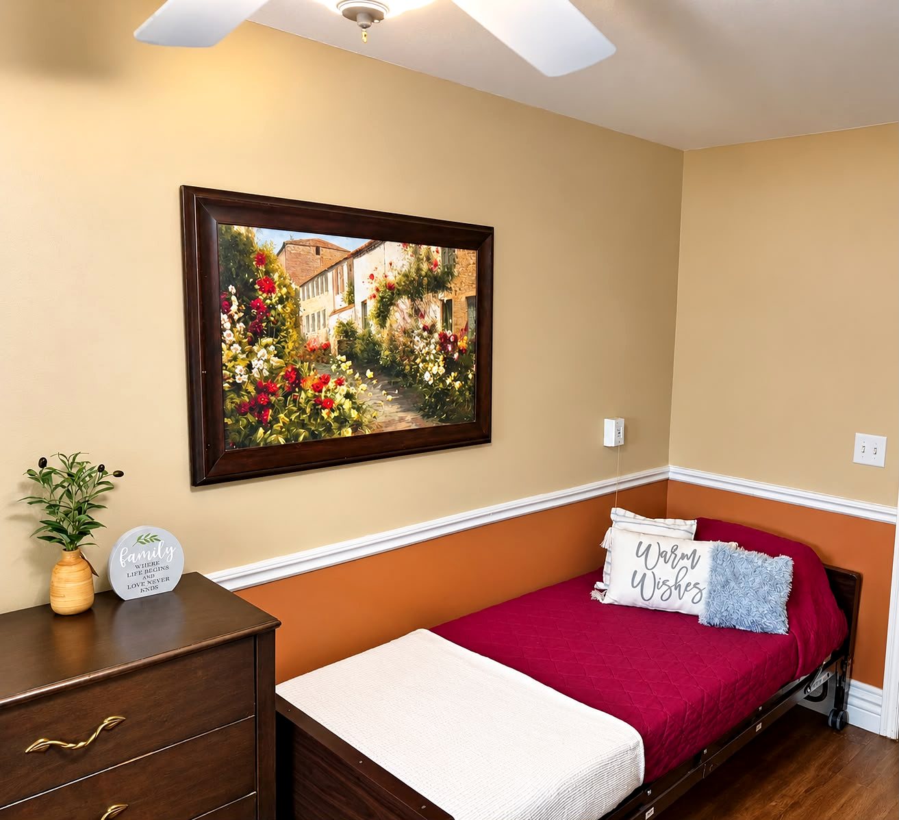 A private bedroom with a red bedspread, a dresser and a framed garden painting